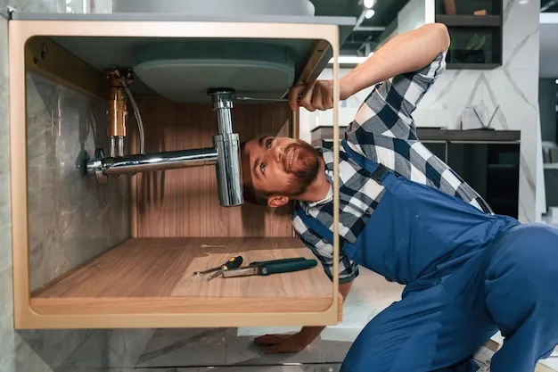 Plumber using wrench on pipes beneath a kitchen sink, surrounded by plumbing tools in a bright home setting.