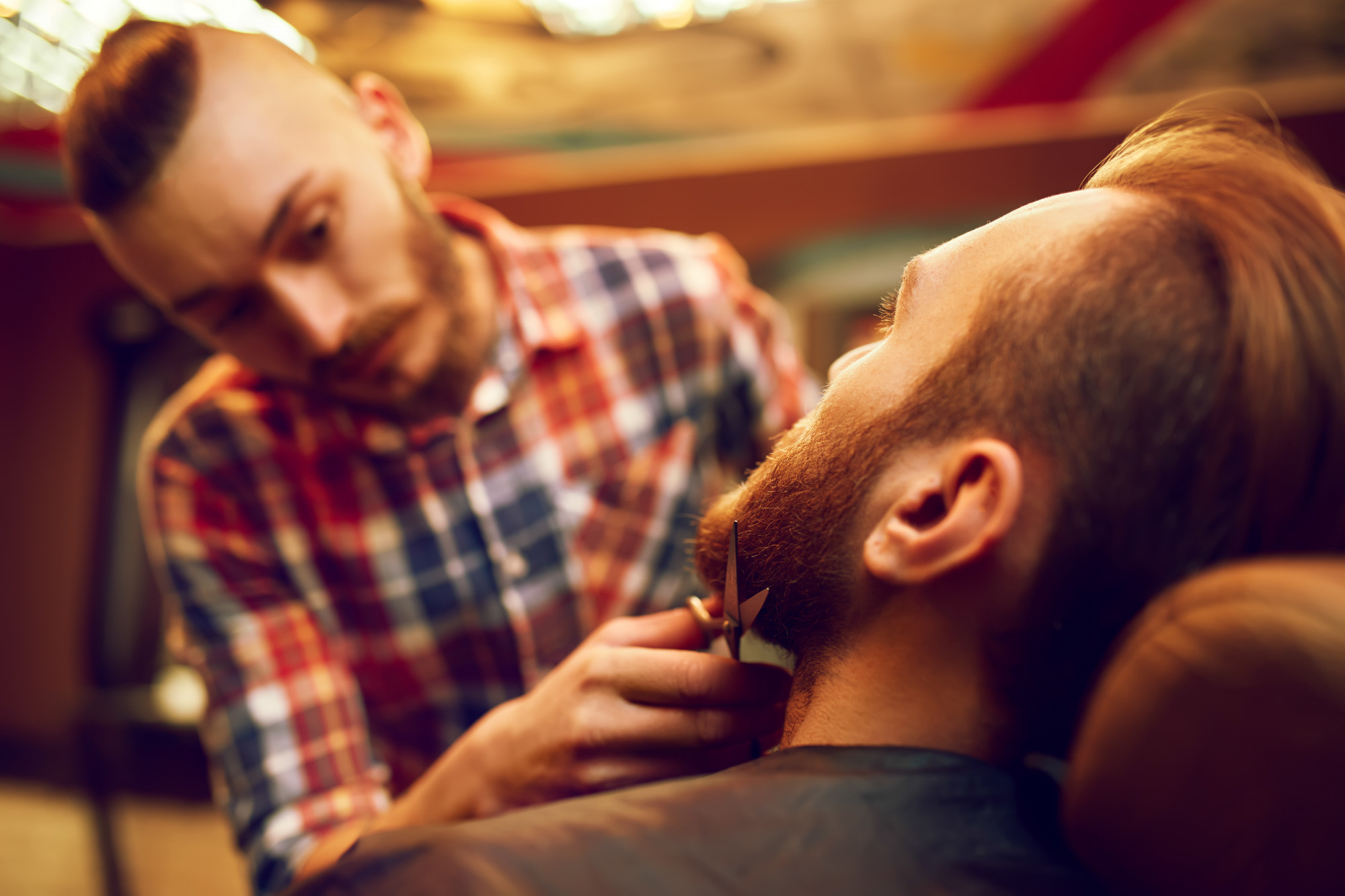 Barber carefully trimming a client's beard with scissors and comb for definition.