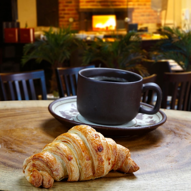 Taza de café con croissant al lado en mesa de panadería.