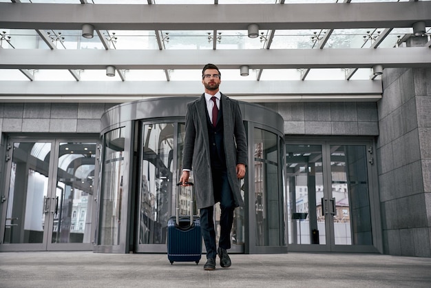 Executive at airport terminal with carry-on, airplane visible through large windows.